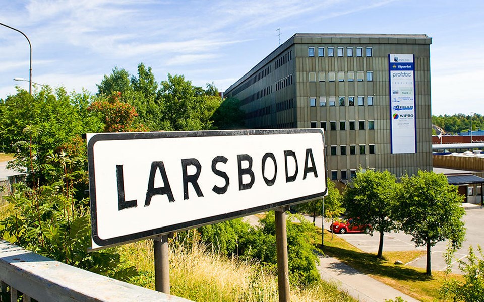 Vy från avfart från Magelungsvägen. Vägmärke med stadsdelsnamnet Larsboda. I bakgrunden flervåningshus, låghus. Parkeringsplats. Foto.