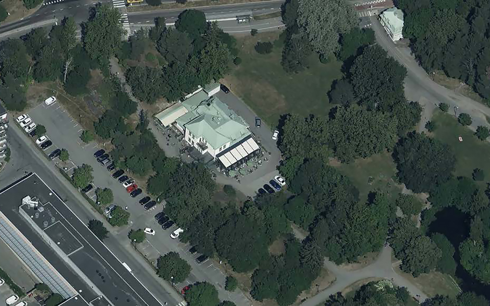 Flygvy över området, byggnad mitt i bild med ljusgrönt tak, parkering, skogsmiljö, fotografi.