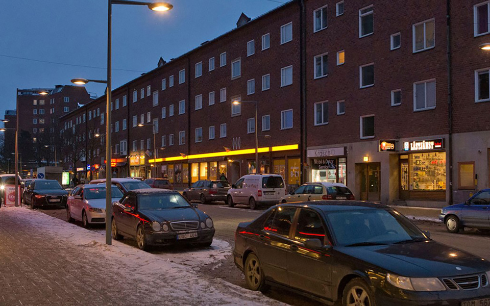 En gata som är upplyst av lyktstolpar. Parkerade bilar på gatan. Upplysta affärer. Människor i rörelse. Foto.