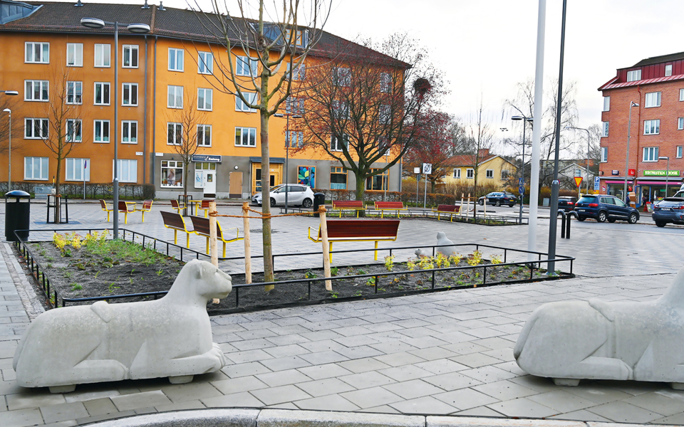 Torg med bänkar och träd omslutet av färgglada flerbostadshus, foto.