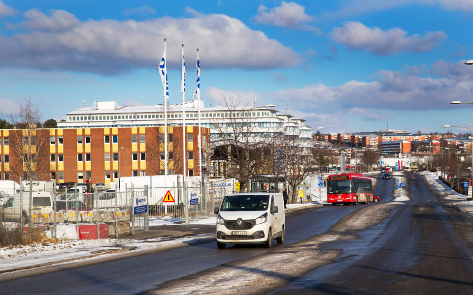 En bilväg med körande bilar. Invid vägen finns industribyggnader och parkering med lastbilar, foto.