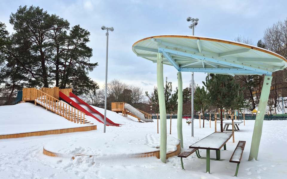 En snötäckt parklek. En parkbänk med tak över syns framför en backe med rutschkanor.