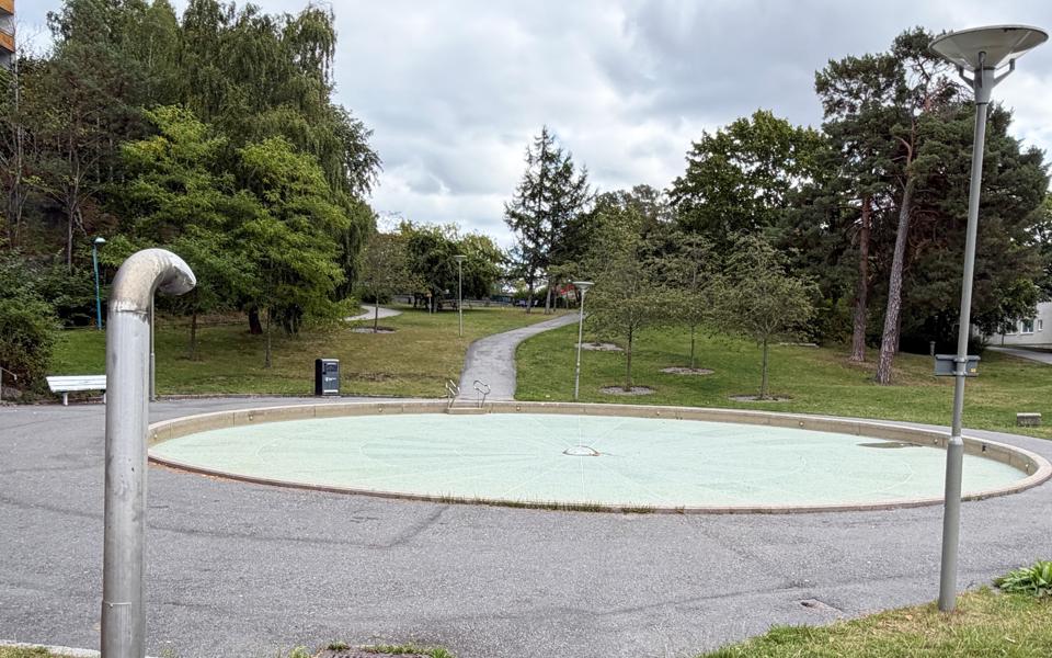 Asfalterad yta med cirkelformad plaskdamm i mitten. Bostadshus, gångväg och träd som omger.