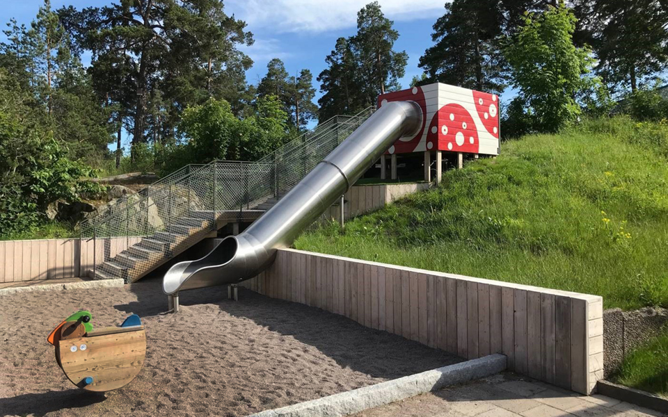 Lekplats med rutschbana, gunghäst och sandlåda, omgiven av gräs och träd. Foto.
