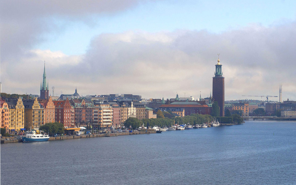 Vy över Norrmälarstrand från vattnet. Båtar förtöjda vid kajen. Bilar på gatan. Flerbostadshus, kyrka och Stadshuset. Foto. 