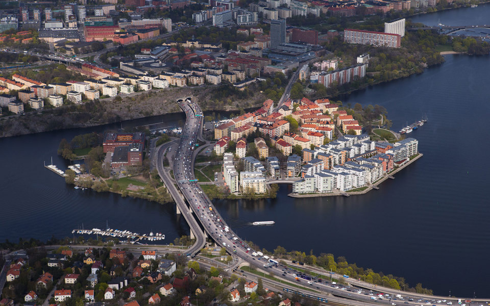 Flygvy över Lilla Essingen. Vägar med rörlig trafik. Bostadsområden och verksamhetsområden. Vatten och båtplatser vid kajer och strandkanter. Foto.