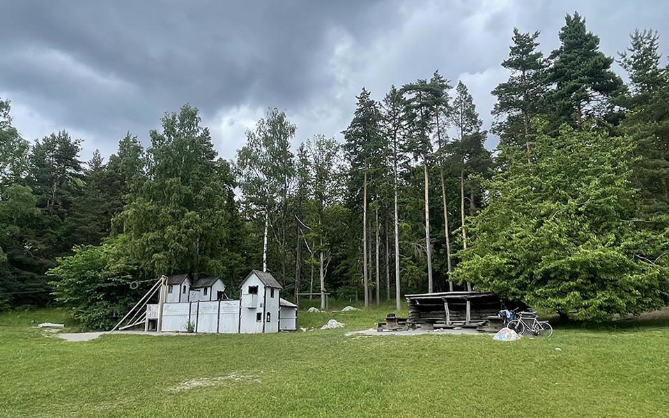 Vy mot lekplats med grillplats, lekställning i form av ett hus och linbana i ett skogsbryn, fotografi.