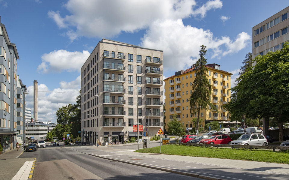 Vy från gata. Flervåningshus med och utan balkonger, parkerade bilar på parkeringsplats, träd, gräs och människor i rörelse, fotografi.