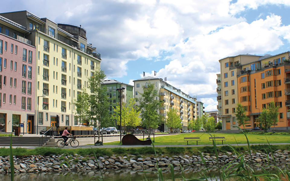 Gräsmatta bakom en vik med flerbostadshus i bakgrunden. En person som cyklar, foto.
