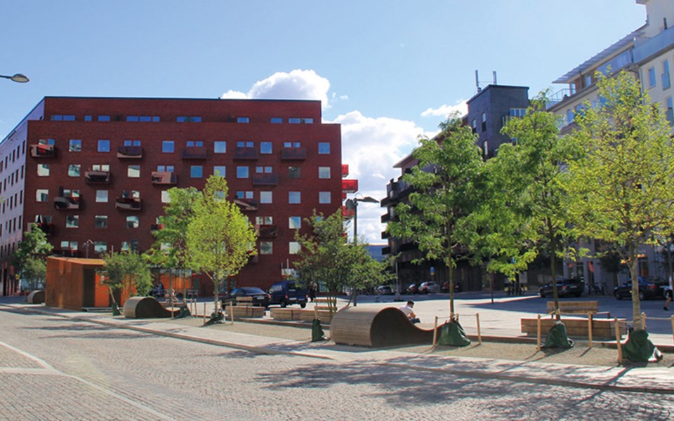 Ett stenbelagt torg med träd och sittplatser,. Omgivet av flerbostadshus, foto.