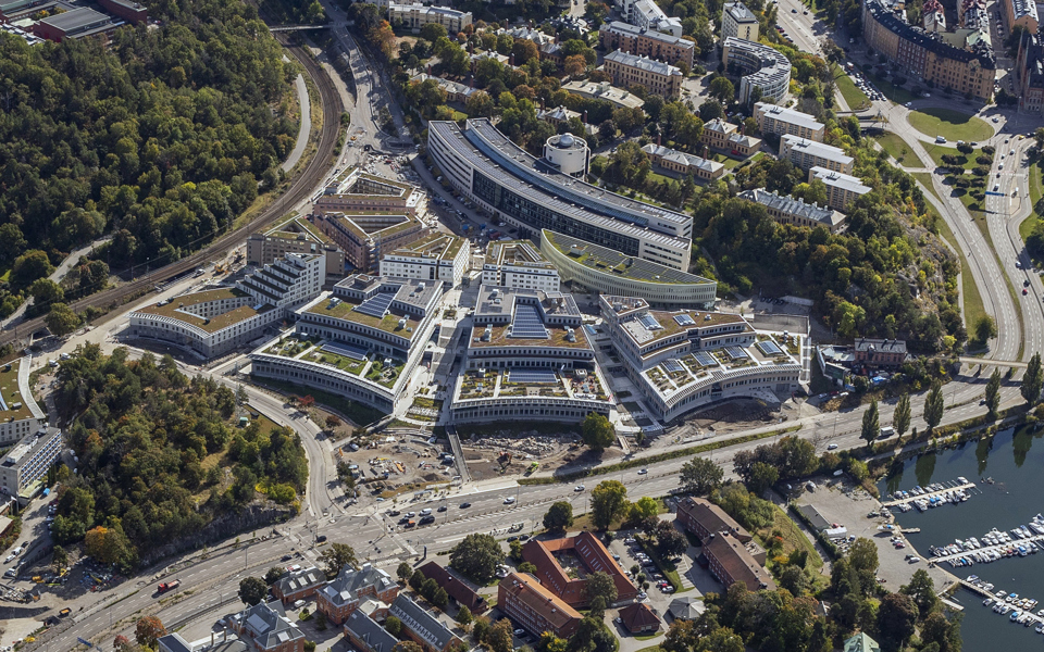 Ett flygfoto där man ser flera stora byggnader i olika nivåer med solceller och växtlighet på taken.