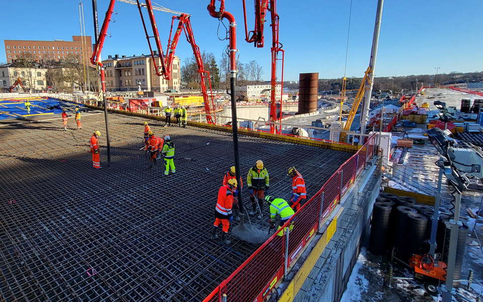 Byggarbetare i arbetskläder gjuter en betonggrund på en stor byggarbetsplats med kranar och utrustning under en klarblå himmel.