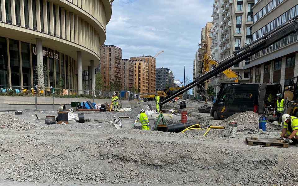 Byggarbetare arbetar på en byggarbetsplats vid Hagaplan i Hagastaden, med flera höghus i bakgrunden och byggutrustning spridd över området.