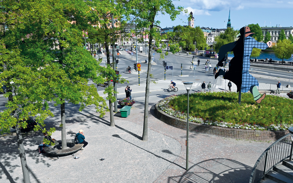 Ett stenbelagt torg med träd och en rund gräsyta i mitten. På gräsytan finns ett konstverk, foto.