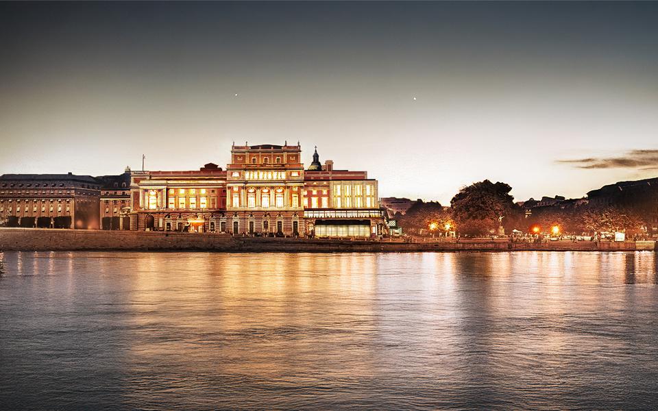 Visualisering av Kungliga Operan i Stockholm med en föreslagen påbyggnad, kvällstid sett från Slottskajen. Byggnaden är upplyst och speglas i vattnet.