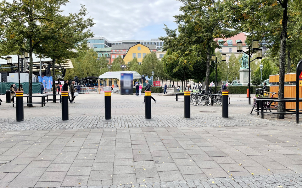Trafikhinder framför torg. I bakgrunden träd,  byggnader, en scen, människor i rörelse, fotografi.
