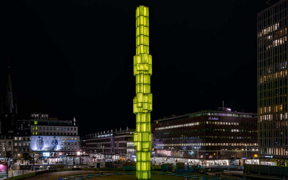 Obelisk upplyst i gult i mitten av en fontän, kväll. Byggnader och neonskyltar i bakgrunden, storstadsmiljö, fotografi.
