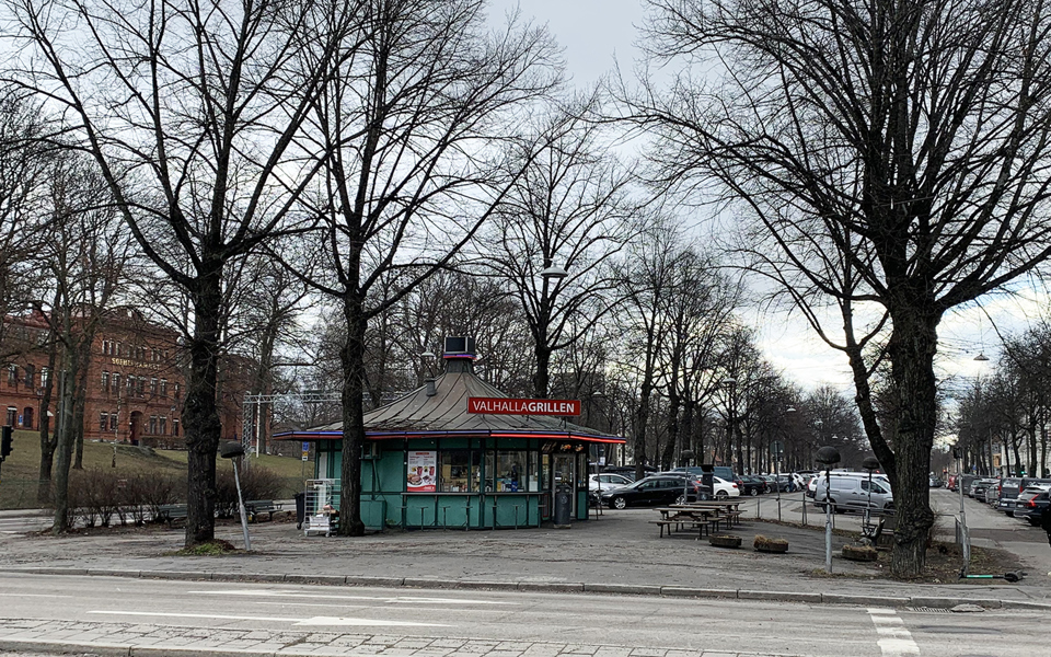Vy i allé mot grillkiosk, med träd utan löv, parkering och byggnad i bakgrunden, fotografi.