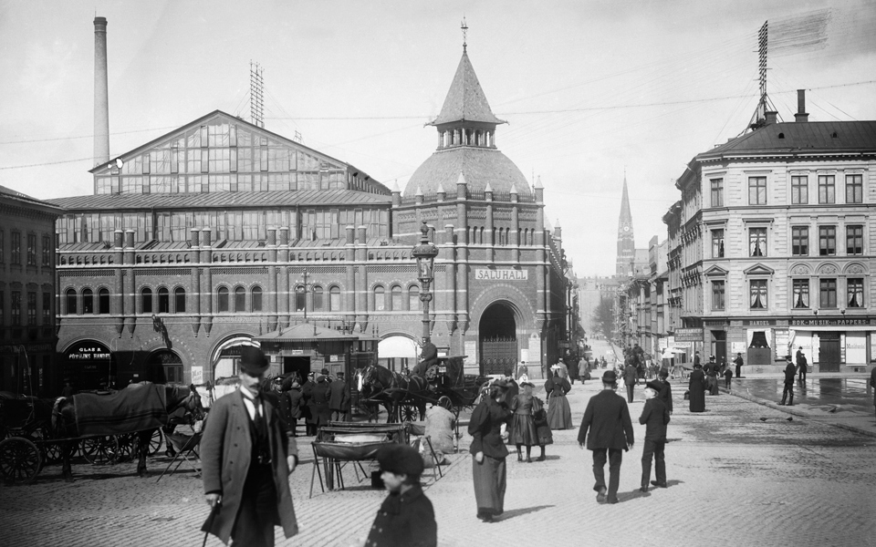 Östermalms saluhall i bakgrunden.  I förgrunden rör sig 1890-talets stockholmare., foto.