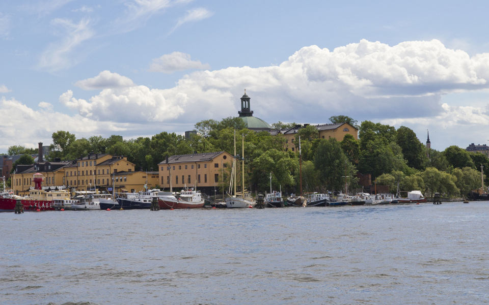 Båtar, byggnader och träd under en blå, molnig himmel. Vy från vattnet.