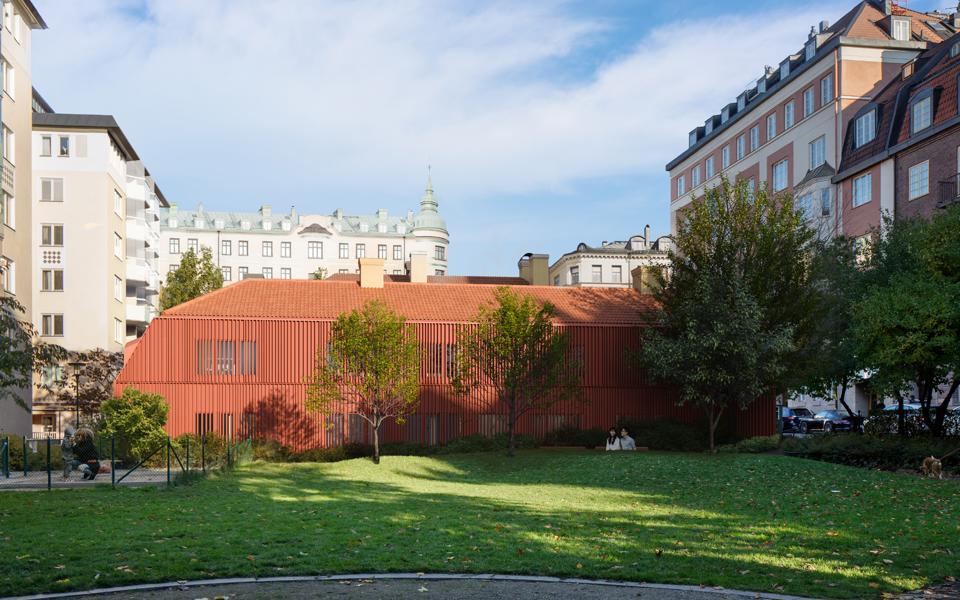 Den tänkta flygelbyggnaden i faluröd panel. Sedd från Monica Zetterlunds park med gräs och träd i förgrunden.