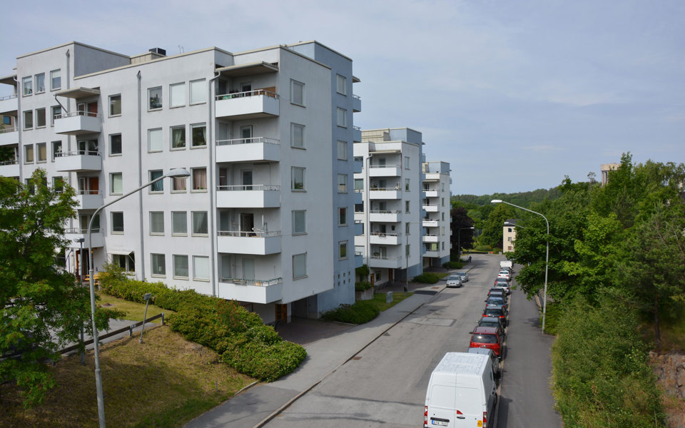Bostadsområde med vita flervåningshus. Väg med parkerade bilar och trottoar i förgrunden.