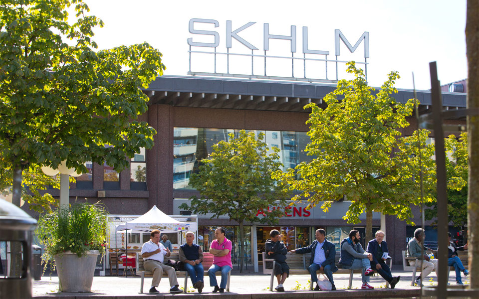 Torg med människor på bänkar. Byggnad i bakgrunden med stor skylt med versalerna "SKHLM" på taket.