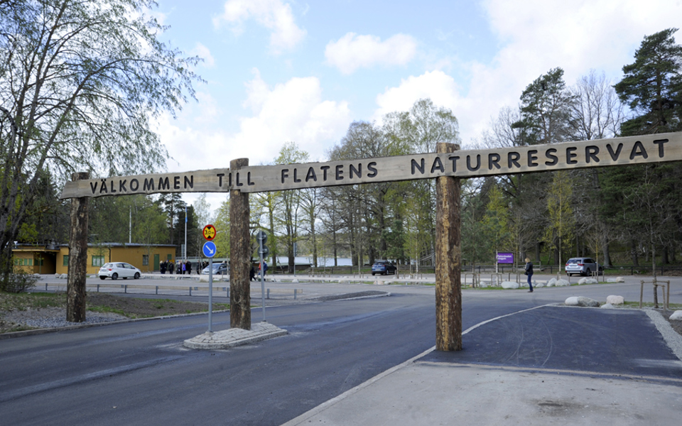 Infart till parkering vid naturreservat. Skylt i trä ovanför infarten  där det står "Välkommen till Flatens naturreservat".