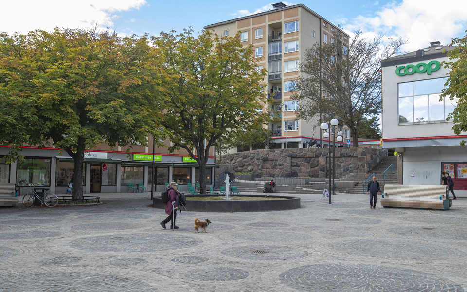 Torg, butiker, flerbostadshus, träd, människor i rörelse, fotografi.