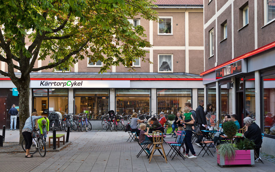 Torg med butik och restaurang. Träd, bordar och stolar. Människor som äter på restaurangen. Cykelställ med cyklar och människor i rörelse. Foto. 