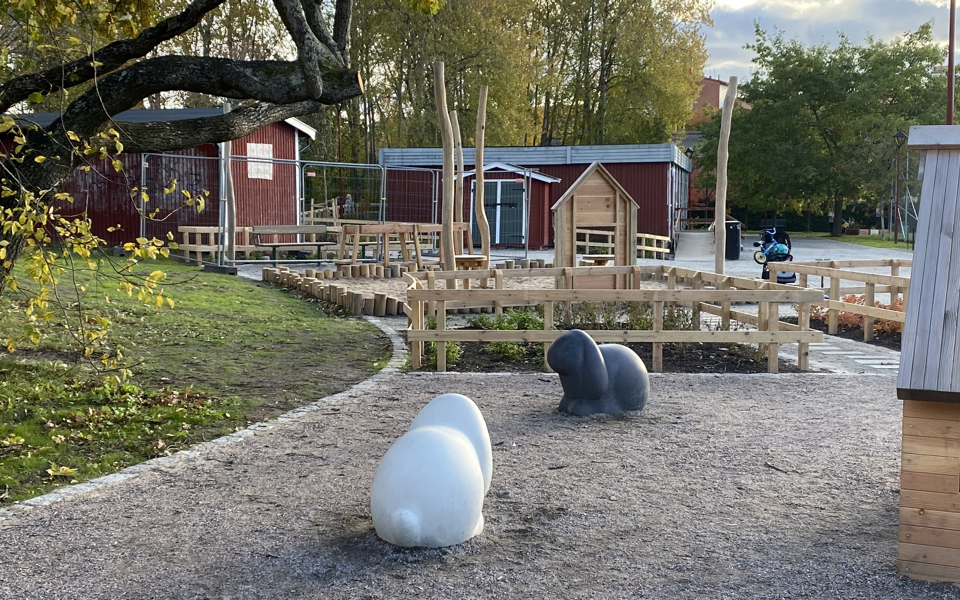 Lekplats med lekhus, kaninstatyer, gångbro i träd, staket runt planteringar, sandlåda, stora träd, sittplatser och en gångväg fram till ett litet rött hus. Foto