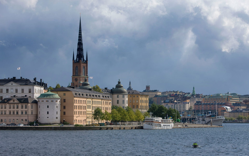 Holme med solbelysta hus och spetsigt kyrktorn bakom. I bakgrunden syns regntunga moln.