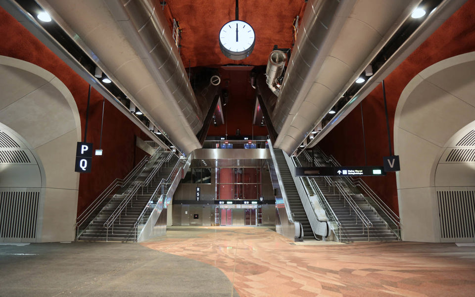 Vy upp mot trappor och rulltrappor i en vänthallsmiljö med rött golv och röda väggar. På båda sidorna framför trapporna finns valv med entréer.