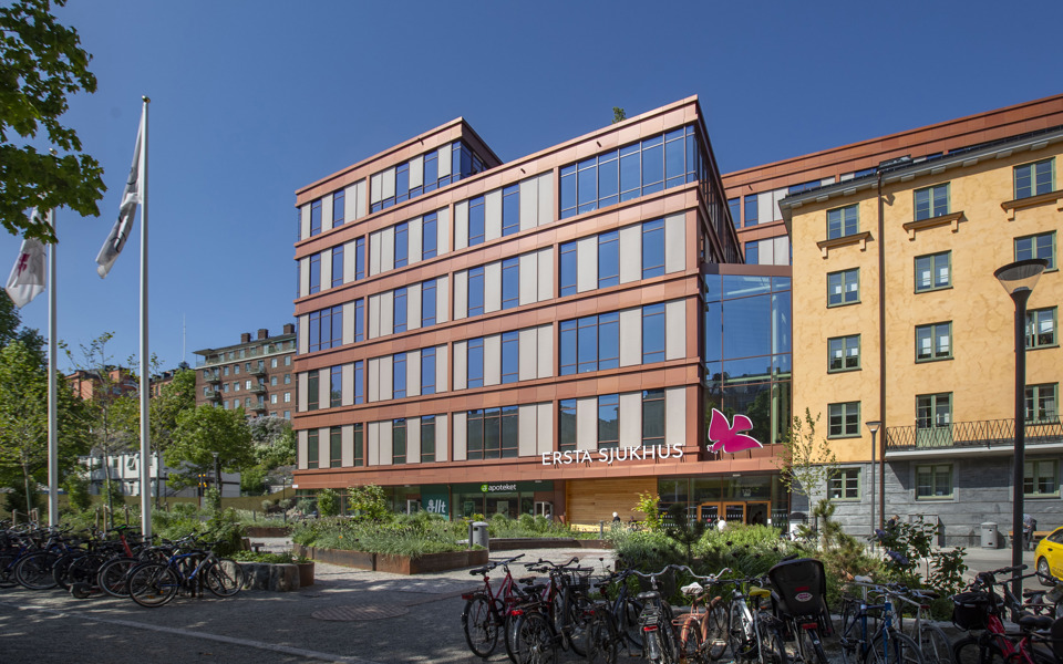 Vy mot byggnad med indragna våningar, stora fönsterglas och fasad med kopparplåt. Ovanför entrén står det Ersta sjukhus och en rosa logotyp. Framför byggnaden finns cykelställ, planterade rabatter och flaggstänger, fotografi.