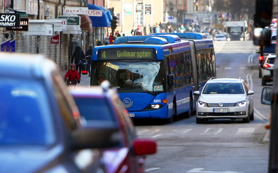 Blå buss som kör fram längs trafikerad gata. Foto.