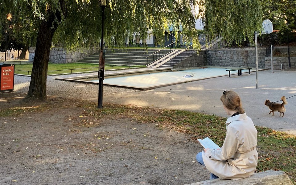 Parkområde med plaskdamm och träd.  En kvinna sitter och läser, foto.