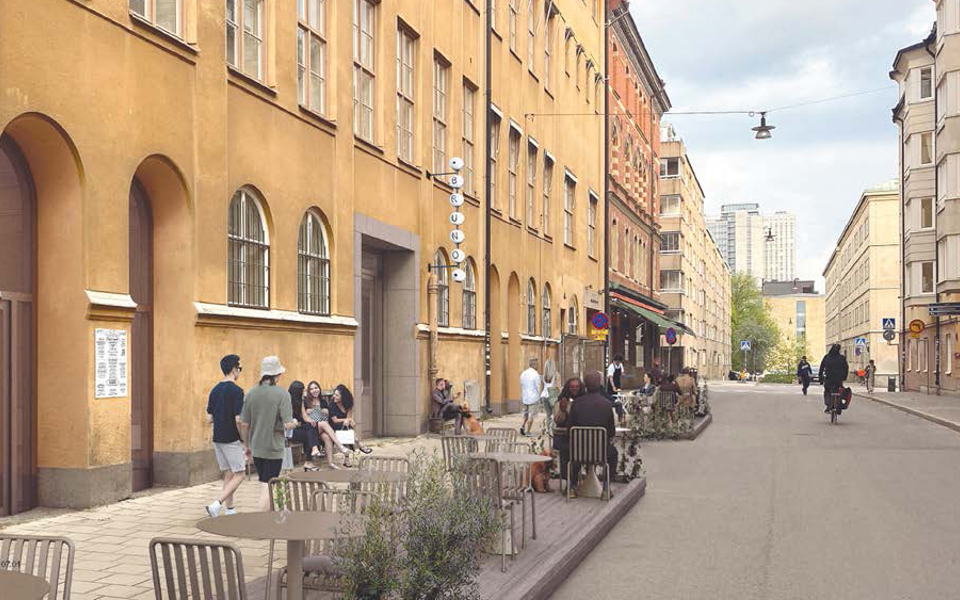 En husfasad vid en gata i innerstaden. Det finns stolar och bord placerade på trottoaren och människor går förbi. 