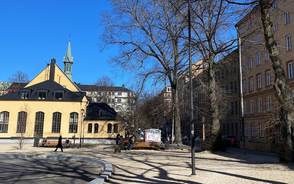 Parkyta omsluten av hus och träd. Några människor som sitter på bänkar och några som promenerar, foto.