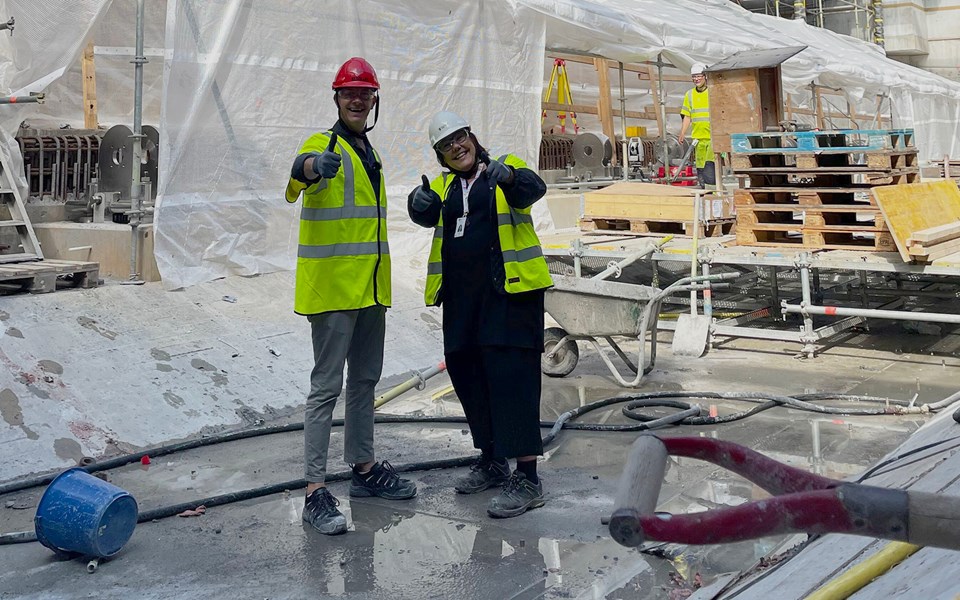 Två personer iklädda gula varselvästar och bygghjälmar står inne på en byggarbetsplats. De är glada och gör tummen upp mot kameran.