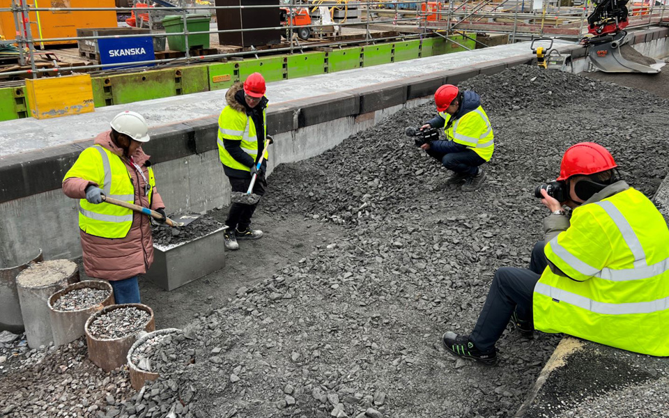 Två personer med gula västar och bygghjälmar står inne på byggarbetsplats, på varsin sida om en låda av plåt. Personerna håller i varsin spade och lägger grus ovanpå lådan. Framför de två personerna och lådan sitter två personer, också iklädda gula västar och bygghjälmar. De fotar händelsen.