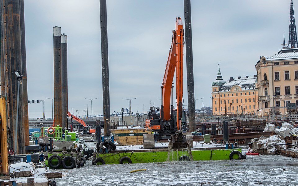 En orange grävmaskin står på en grön ponton på vatten och gräver. I bakgrunden ligger Gamla stan.