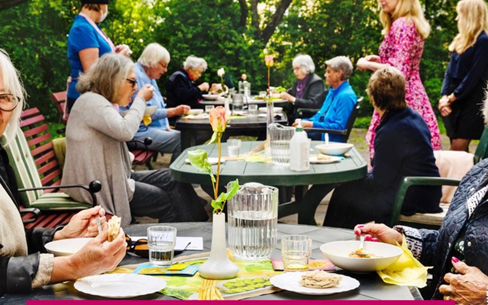 Två äldre personer sitter i en trädgård, andra människor syns i bakgrunden