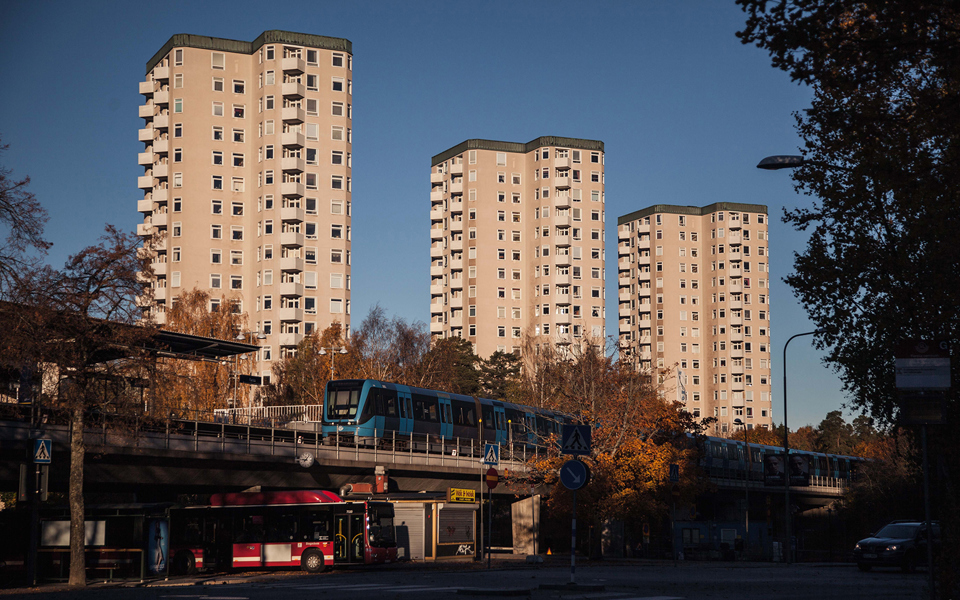 Ett tunnelbandetåg åker in på perrongen som ligger  ovanför en busstation. I bakgrunden syns tre höga flervåningshus.