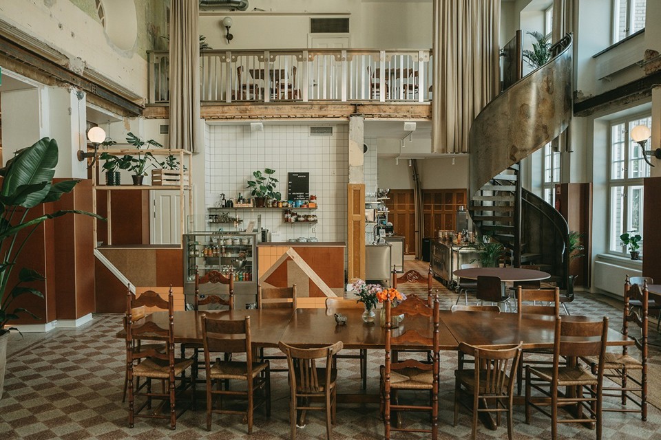 Färgbild av Börssalen i Börshuset, inrett som ett kafé med träbord och stolar. En spiraltrappa finns vid väggen till höger och en kaffebar syns i bakgrunden. Stora fönster släpper in naturligt ljus, och gröna växter finns i rummet.