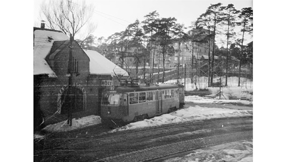 En historisk svartvit bild från 1950-talet som visar en spårvagn framför en tegelbyggnad med skylten "Kött". Marken är täckt av snö och i bakgrunden syns träd och flerfamiljshus på en sluttning.