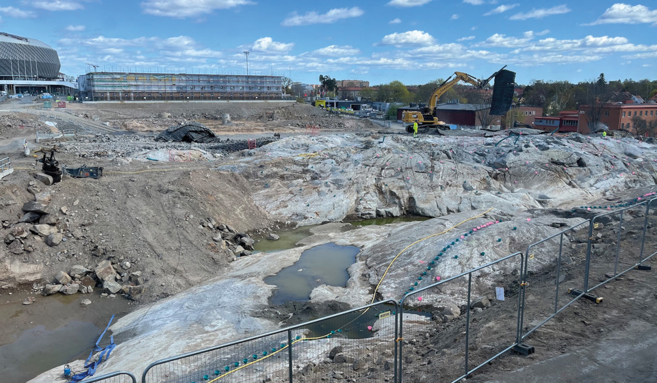 En byggarbetsplats med grävmaskiner, stängsel och ett stenigt landskap under konstruktion, med en stor arena och byggnader i bakgrunden.