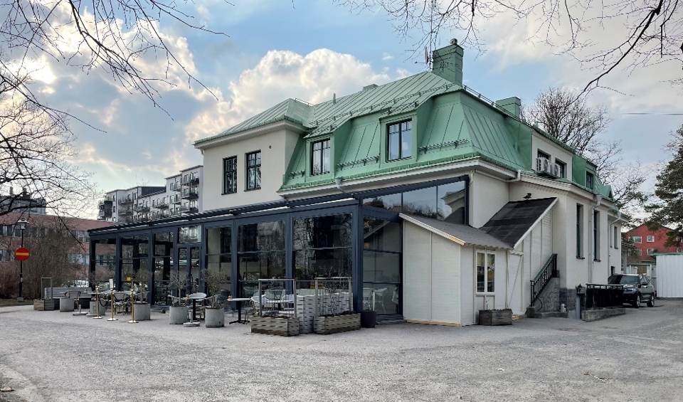 Byggnad med putsad fasad och ljusgrönt plåttak med tillbyggnad i glas. Framför tillbyggnaden finns en uteservering, fotografi.