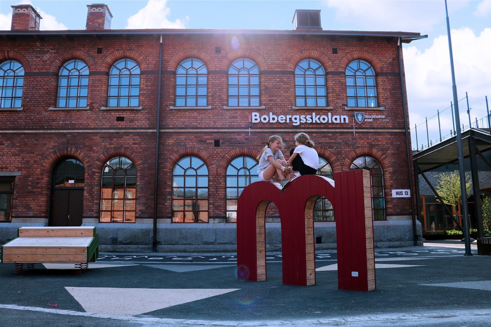 Två barn som sitter på en skulptör som är format som ett m på skolgården. Foto.